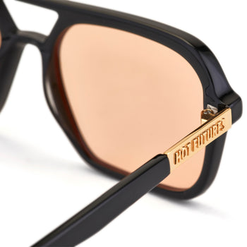 A pair of slim rectangular aviator sunglasses with a black frame and amber tinted lenses. With gold Hot Futures branding text shown on the arms. 