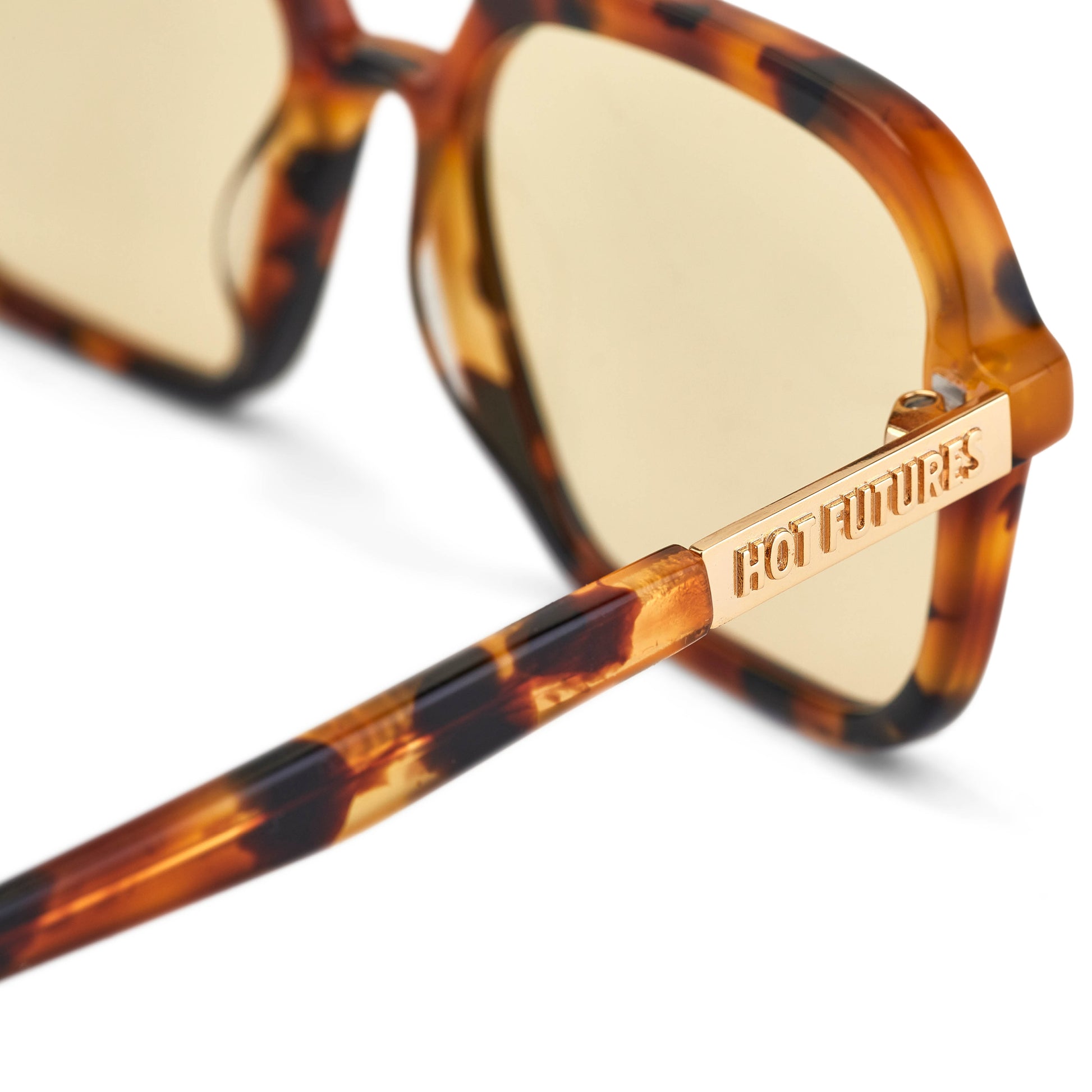 A pair of slim rectangular aviator sunglasses with a classic tortoiseshell pattern and gold tinted lenses. Shot close up showing gold Hot Futures branding text