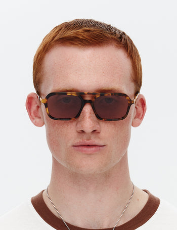Model wearing a pair of tortoiseshell patterned sunglasses with a slim rectangular aviator frame 